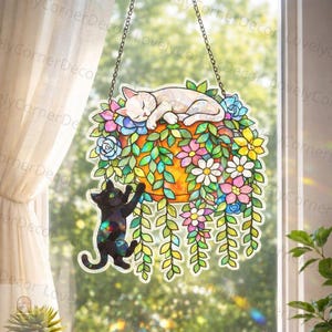May include: Stained glass window hanging depicting a sleeping white cat on a floral arrangement with a black cat climbing below. The arrangement includes colourful flowers and green foliage, suspended by a chain.