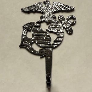 May include: A dark metal wall hook featuring the emblem of the United States Marine Corps. The emblem includes an eagle perched atop a globe, an anchor, and a stylized design. The hook is suitable for hanging items.