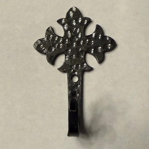 May include: A dark metal wall hook with a fleur-de-lis design. The hook has a hammered texture and a small loop for hanging. The hook is a decorative item for home decor.