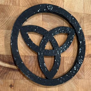May include: A black metal trivet with a Celtic knot design. The trivet has a circular frame and a three-part knot in the centre. The metal has a textured, speckled appearance. It is designed to protect surfaces from hot cookware.