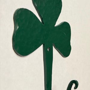 May include: A green metal shamrock hook with a textured finish. The hook is in the shape of a three-leaf clover with a curved hook at the bottom. The hook is designed for hanging items and is suitable for St. Patrick's Day decor.