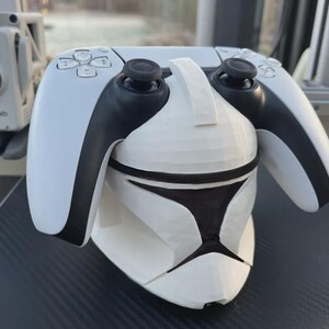 Może przedstawiać: Biało-czarny kontroler do gier spoczywający na stojaku w kształcie hełmu żołnierza-klona z Gwiezdnych Wojen. Kontroler ma czarne joysticki i przyciski. Stojak jest biały z czarnymi akcentami.