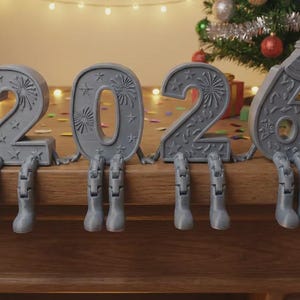 Könnte beinhalten: Graue Tischdekoration für Silvester 2026 mit dekorativen Feuerwerks- und Sterndesigns. Die Zahlen haben bewegliche Beine und sitzen auf einer Holzoberfläche. Ein Weihnachtsbaum und Lichter befinden sich im Hintergrund.