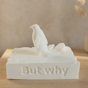 May include: A white penguin ornament standing on a rectangular base. The base features the words "But why". The sculpture is placed on a wooden surface, with a neutral backdrop.
