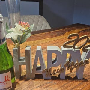 May include: A green bottle with a white label and a silver cap sits next to a gray and gold "Happy New Year 2026" decoration. A small vase with white and pink flowers is also present.