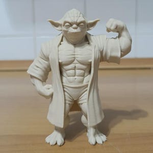Può includere: Statuetta beige di un personaggio Yoda muscoloso che flette il braccio. La figura indossa una veste e pantaloncini. La statuetta ha grandi orecchie e un'espressione severa. La figura è su una superficie di legno.