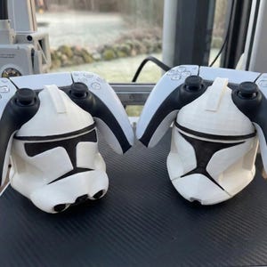 Może przedstawiać: Dwa biało-czarne kontrolery PlayStation 5 spoczywają nad pasującymi białymi i czarnymi uchwytami w kształcie hełmu Clone Troopera. Hełmy mają czarne oznaczenia twarzy.
