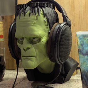 Può includere: Un busto della testa di Frankenstein verde con cuffie nere. Il busto ha una base nera e si trova su una superficie di colore chiaro. Una bottiglia di soda e una tazza con un design colorato sono sullo sfondo.