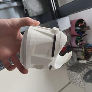 Może przedstawiać: Biały, drukowany w 3D obiekt o złożonym geometrycznym wzorze, trzymany w dłoni. Obiekt ma zakrzywioną, otwartą sekcję i jest zabezpieczony czarnymi paskami. Tło obejmuje drukarkę 3D i r&oacute;żne szpule.