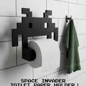 May include: A black toilet paper holder shaped like a pixelated alien character from a video game. The holder is mounted on a wall and holds a roll of white toilet paper. The text SPACE INVADER TOILET PAPER HOLDER | RETRO ARCADE DECOR is visible.