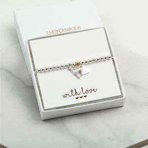 Peut inclure: Un bracelet à breloques en argent avec un petit charme en forme de cœur en argent et un charme en argent représentant un chien avec un accent doré. Le bracelet est dans une boîte blanche avec les mots "LHO Designs" et "with love" imprimés dessus.