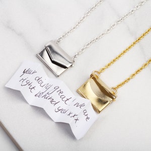 May include: Two silver and gold necklaces with small envelope pendants. The necklaces are on a white marble surface. A handwritten note reads "Your doing great! We are right behind you xxx."