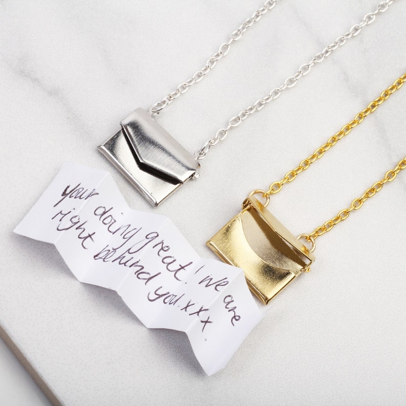 May include: Two silver and gold necklaces with small envelope pendants. The necklaces are on a white marble surface. A handwritten note reads "Your doing great! We are right behind you xxx."