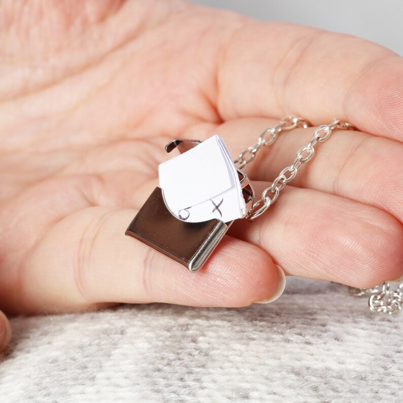 May include: A silver locket necklace with a small, rectangular, hinged box. The box is open and contains a folded piece of paper with the letters 'O' and 'X' written on it.