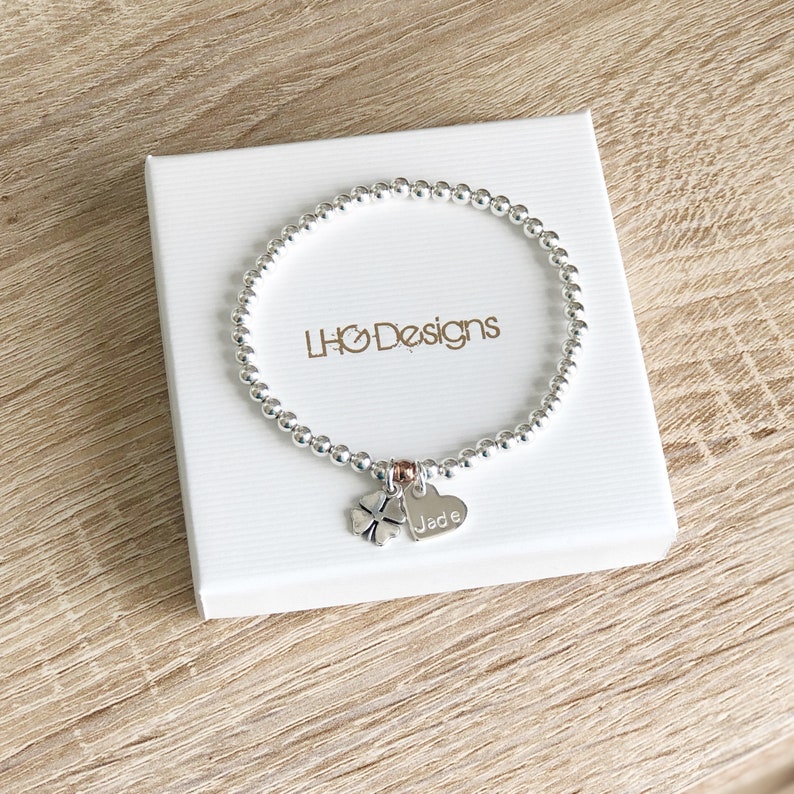 Pu&ograve; includere: Un braccialetto in argento con perline, un ciondolo a forma di quadrifoglio in argento e un ciondolo a forma di cuore in argento con inciso il nome "Jade". Il braccialetto &egrave; in una scatola bianca con la scritta "LHO Designs" stampata.