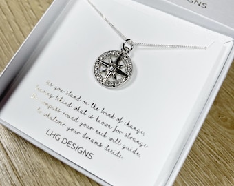 May include: A silver compass pendant necklace displayed in a white gift box. The compass pendant is round, with a star design and encrusted with small, clear crystals. The box contains text that reads "As you stand on the brink of change..." and the brand name "LHG DESIGNS".