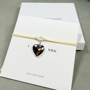 May include: A silver heart charm bracelet with the words "I YOU" printed on a white card. The card is on top of a white box with the word "DESIGNS" printed on it. The bracelet is on a cream-colored cord.