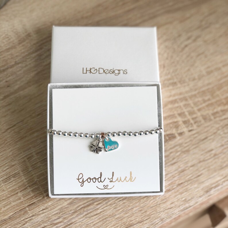 Pu&ograve; includere: Un braccialetto d'argento con un piccolo ciondolo a quattro foglie di trifoglio d'argento e un ciondolo a forma di cuore turchese con il nome "Jade" inciso su di esso. Il braccialetto &egrave; in una scatola bianca con le parole "LHG Designs" e "Buona fortuna" stampate su di essa.