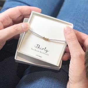 May include: A white box containing a silver and gold beaded bracelet. The box has the text "Thirty" and "Three Beads Three Decades" printed on it.