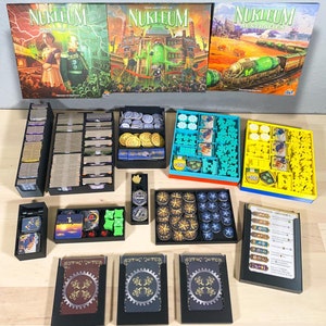 May include: A collection of black plastic storage boxes filled with game pieces and cards for the board game "Nukleum". The game boxes are arranged on a wooden table. The game boxes are labelled with the game title "Nukleum" and the game expansions "Overseas" and "Australia".