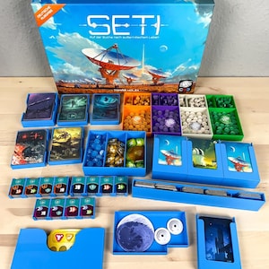 May include: A blue box with the text "SETI" and the words "Ruf der Suche nach außerirdischem Leben" on the front. The box is open and contains various game components, including cards, dice, and tokens. The components are organised in blue plastic trays.