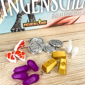 Könnte beinhalten: Spielsteine, darunter Goldbarren, lila Kristalle und silberne Spielmarken. Die Teile sind auf einer Holzoberfläche angeordnet, mit einer Spielbox im Hintergrund. Auf der Box steht "INGENSCHLAG" und "FEUERLAND".