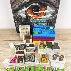 May include: A board game box with the word "RECAL" on top, featuring an eye illustration. Various game components are arranged in white and coloured trays, including cards, tokens, and dice. The game pieces are in various colours, including green, yellow, and pink.