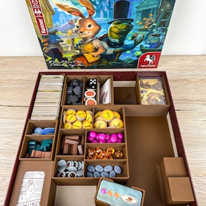 Può includere: Scatola di gioco da tavolo aperta "Everdell Duo" con un'illustrazione di coniglio e tartaruga. La scatola contiene componenti di gioco, tra cui carte, dadi e gettoni colorati. I pezzi sono organizzati in scomparti marroni.
