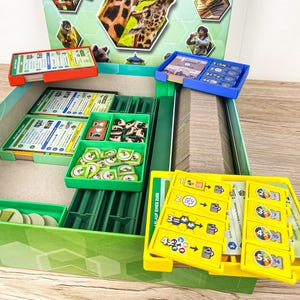 May include: A board game box with the title "Arten Garten" and the subtitle "Ein Arche Nova-Spiel". The box features images of giraffes, a toucan, and other animals. Inside, there are various game components, including cards and tokens, organized in green, red, and blue trays.