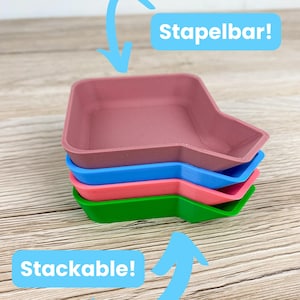 Peut inclure: Une pile de quatre plateaux empilables colorés, verts, roses, bleus et d'une couleur rose clair. Les plateaux sont rectangulaires avec un design angulaire unique. Le texte sur l'image indique "Stackable!" et "Stapelbar!"