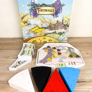 May include: A board game titled Fromage featuring a box with a landscape illustration, a white plastic tray with game cards and tokens, a hexagonal game board with small cheese-shaped markers, and four large wedge-shaped cheese pieces in white, black, red, and blue.
