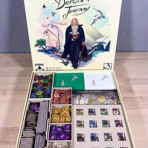 Può includere: Una scatola beige con un inserto di gioco per il gioco da tavolo "Darwin's Journey". L'inserto ha scomparti per vari componenti di gioco, tra cui gettoni, carte e tessere. Il coperchio della scatola presenta un'illustrazione di Charles Darwin su una nave, con il titolo del gioco "Darwin's Journey" scritto in corsivo.
