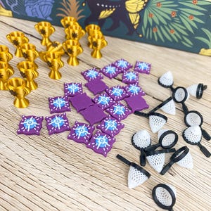 Puede incluir: Componentes de juego de mesa: trofeos dorados, fichas cuadradas moradas con diseños blancos y azules, y pequeñas redes blancas y negras. Las piezas están dispuestas sobre una superficie de madera clara.