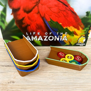 May include: Board game components featuring brown boat-shaped containers with colorful trim and game tokens. The background shows a vibrant parrot illustration and the text "LIFE OF THE AMAZONIA".