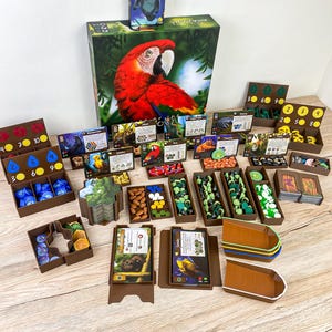 Può includere: Un set di gioco da tavolo con una scatola grande raffigurante un ara rossa, una scatola più piccola con un lamantino e numerosi organizzatori in plastica marrone pieni di carte da gioco, gettoni e pezzi in legno. Include varie carte animali.