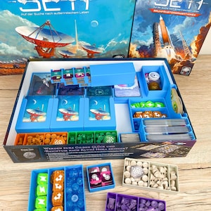 Puede incluir: Juego de mesa "SETI" con temática de exploración espacial. La caja presenta ilustraciones de una antena parabólica y cohetes. En el interior, hay varias piezas del juego, cartas, fichas y dados, organizadas en bandejas azules y de colores.
