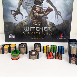 Puede incluir: Un juego de mesa titulado "The Witcher: Die Alte Welt" con varias piezas. Las piezas del juego están en soportes negros e incluyen pilas de monedas y fichas de colores. La caja del juego presenta una ilustración de fantasía con lobos y un guerrero.