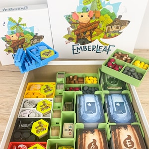 Könnte beinhalten: Emberleaf-Brettspiel mit einer Schachtel mit einer Mausfigur, die Holz hält, einer Windmühle und dem Spieletitel. Die offene Spielbox zeigt verschiedene Spielsteine, Karten und Aufbewahrungsfächer in Grün-, Gelb- und Blautönen.