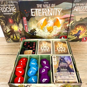 May include: The board game The Vale of Eternity with two expansions, Fluche and Artefakte, displayed behind an open box containing red, blue, and purple tokens, black cubes, and a deck of cards featuring the character Tolvar der Bodrückte.