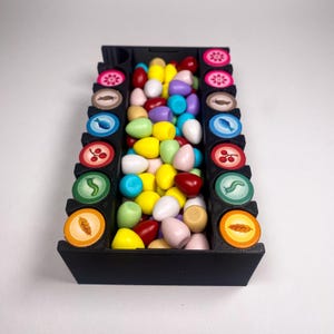 May include: A black plastic storage box with two rows of colorful egg-shaped tokens. The box has a variety of symbols on the front, including a leaf, a snail, a berry, and a fish.