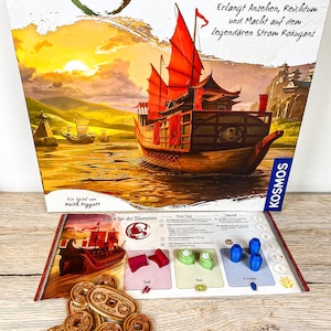 Könnte beinhalten: Brettspiel "Am Goldenen Fluss" mit einer Schachtel, die ein Gemälde eines rot besegelten Schiffs auf einem Fluss zeigt. Spielkomponenten sind Holz-Münzen, Spielsteine und Karten. Das Spiel ist von Keith Piggott.