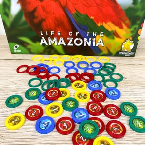 Könnte beinhalten: Brettspiel "Life of the Amazonia" mit einer Illustration eines roten Papageis. Das Spiel enthält bunte Spielsteine und Ringe in Rot, Blau, Grün und Gelb. Die Spielsteine haben Zahlen und Blattillustrationen.