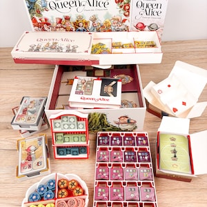 May include: A Queen Alice board game set featuring a large game box, a smaller box, card decks, a white plastic tray with four compartments containing blue, yellow, orange, and pink flower tokens, and a rectangular tray with pink tiles.
