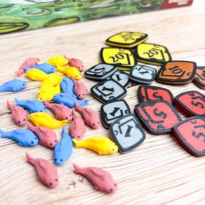 Peut inclure: Une collection de petits jetons poissons en plastique rose, jaune et bleu, accompagnés de tuiles de jeu carrées jaunes, grises, rouges et brunes. Les tuiles présentent des chiffres noirs imprimés dont 1, 5, 10 et 20. Une boîte de jeu est visible.