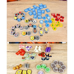 Puede incluir: Una colección de componentes de juego de mesa que incluye varios fichas hexagonales y circulares en azul, rojo, amarillo, verde, blanco y morado. El set presenta pequeñas piezas de recursos de plástico, losetas numeradas y piezas de panal.