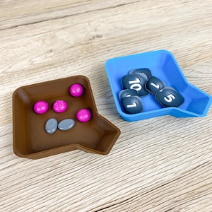Peut inclure: Deux petits plateaux ouverts reposent sur une surface en bois. L'un est marron et contient des pièces de jeu roses et grises. L'autre est bleu et contient des tuiles de jeu noires avec des chiffres blancs. Les plateaux sont conçus pour les jeux de société.