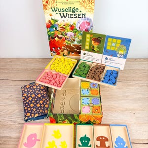 May include: A Wuselige Wiesen board game set featuring a large illustrated box, two character cards for Belota and Siella, and multiple wooden game pieces in yellow, pink, green, brown, and blue. Includes several rectangular trays with colourful icons.