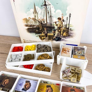 May include: A board game titled KILIA featuring a box with a harbour illustration and multiple white plastic trays containing game components. The trays hold red and yellow wooden cubes, grey and gold coins, and various cards with hand-shaking graphics.