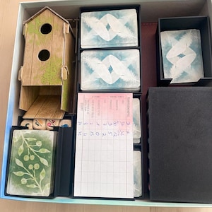 Op de afbeelding: Een speldoos met een houten vogelhuisje, een scoreblad en verschillende stapels kaarten. Het scoreblad heeft handgeschreven nummers en de tekst "Mehrpersonenspiel" wat "multiplayer game" betekent.