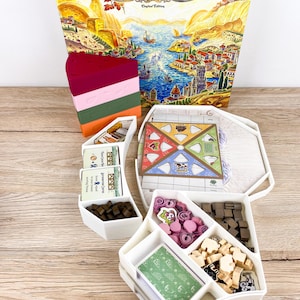 Może przedstawiać: Pudełko gry planszowej Formaggio Limited Edition zawiera ilustrację krajobrazu przybrzeżnego. W zestawie znajduje się stos czterech kolorowych klinów w kształcie sera oraz biała plastikowa tacka z taliami kart, małymi brązowymi kostkami, różowymi żetonami i beżowymi figurkami zwierząt.